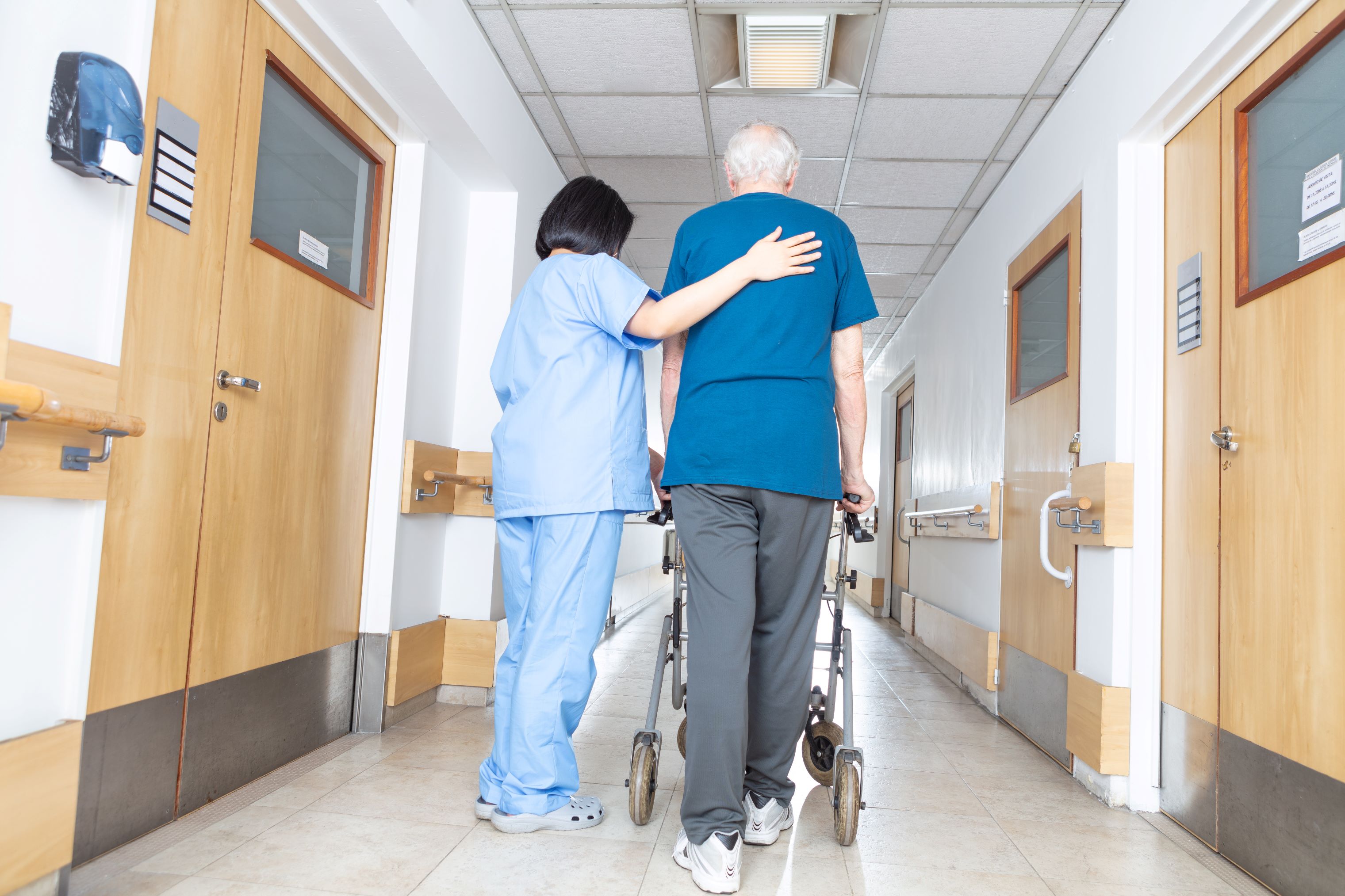 Nurse assists senior man using a walker down hallway in care facility.